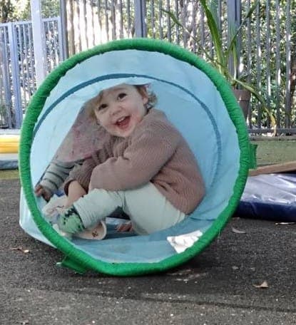 A smiling toddler playing in a tunnel in the centre's outdoor play area