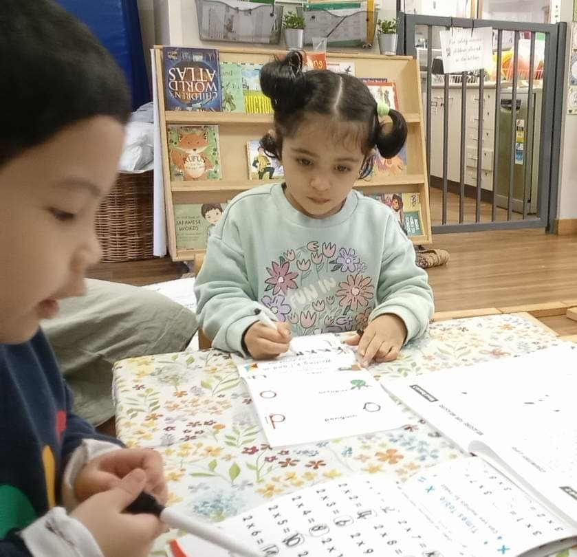 Two kindergarten children practising early writing, surrounded by multicultural picture books